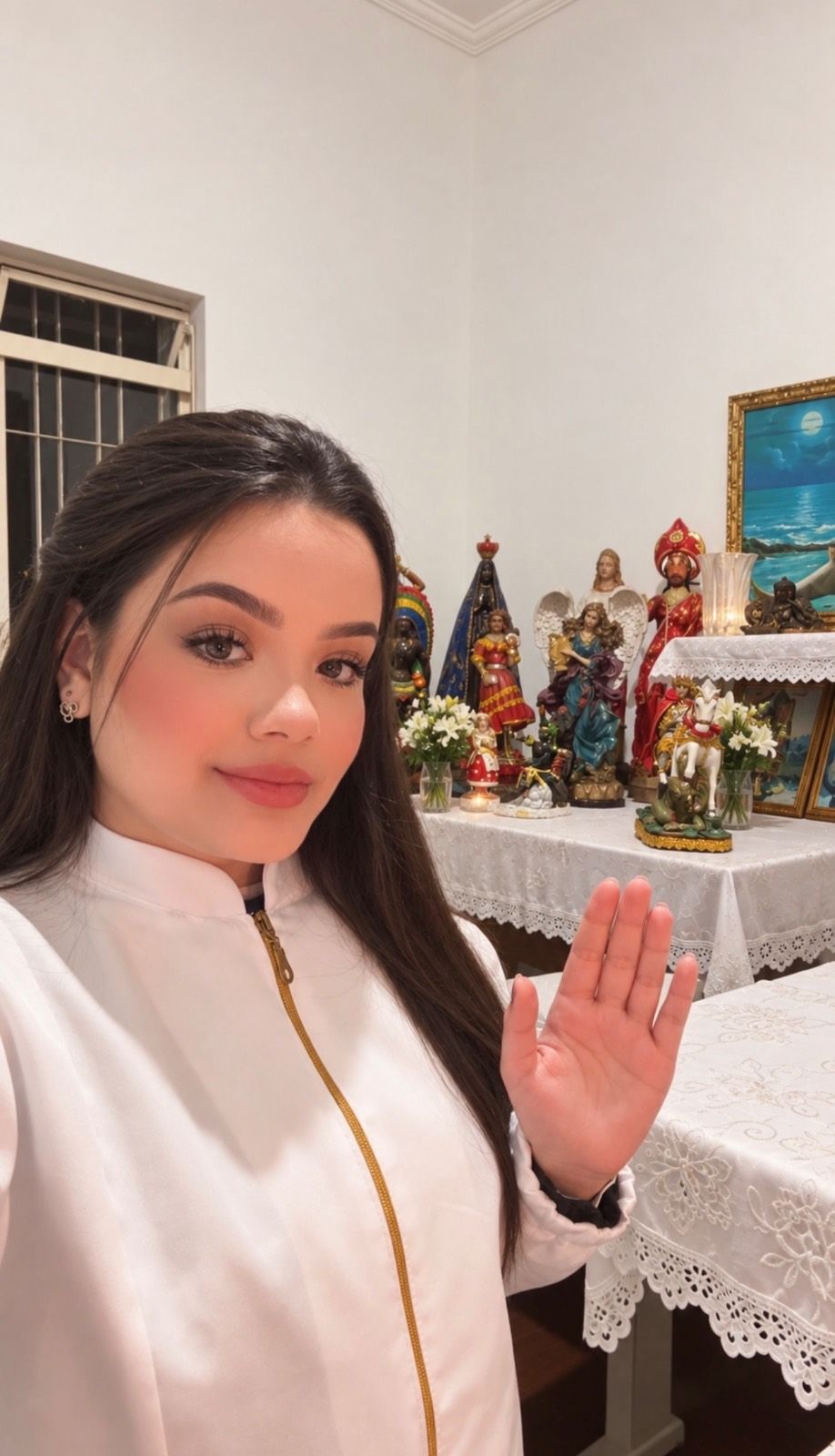 Vidente Samyra de jaleco branco acenando em frente ao altar com imagens de santos, orixás e flores brancas